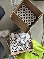 Two open cardboard boxes filled with white receipt paper rolls, some neatly stacked and others scattered.