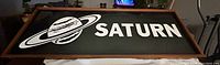 Photo of the full Saturn sign displaying the graphic and logo clearly on a black background within a wooden frame.