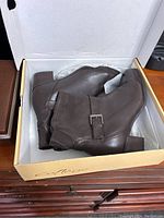 Pair of brown leather boots inside an open box showing the side profile with buckle and block heel detail.