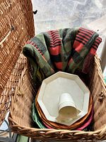 Wicker picnic basket opened showing contents including folded retro-patterned blanket, white plastic octagonal plates, white plastic cups, and multi-colored small woven plates or trays.