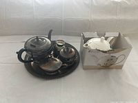 Silver tone metal teapot set with teapot, sugar bowl, creamer, and tray alongside white ceramic teapot in box.