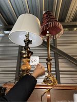 Photo showing two decorative table lamps; one has a white cylindrical shade and mixed material base, the other has a red fringed shade and brass base. Both lamps are placed on a wooden surface with cords visible.