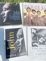 Four Beatles and John Lennon related books laid flat on outdoor surface with greenery background.