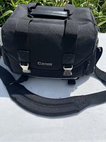Front view of the Canon camera bag showing the flap with Canon logo, metal buckles, padded top handle, and padded shoulder strap.