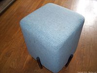 Blue upholstered foot stool, cube-shaped, sitting on a hardwood floor showing the side angle and the black legs.