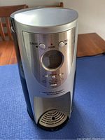 Front view of stainless steel Hamilton Beach BrewStation Brewmaster Deluxe coffee maker on a blue surface, showing control panel with buttons, digital display, and drip tray.