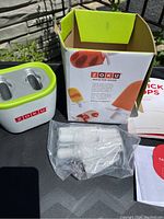 Photo of Zoku Quick Pop Maker device outside its original box with instruction manual and multiple white plastic pop sticks still in plastic wrapping placed next to it on a table.