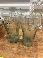 Two Coca Cola glasses, one smooth with vertical grooves, one textured with embossed logos on wooden surface.