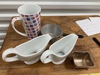 Overview of all five items on table: 2 white ceramic gravy boats, polka dot mug, aluminum measuring cup, and silver square bowl