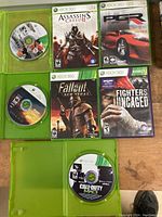 All seven Xbox 360 game discs with cases visible including Assassin's Creed II, Fallout New Vegas, etc.