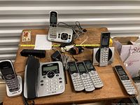 Lot of mixed cordless and corded home phones from Uniden and VTech shown on a wooden surface with some cables and power adapters.
