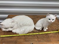 Two white ceramic cat figurines on a wooden surface, one sleeping and one sitting, with a measuring tape showing length and height.