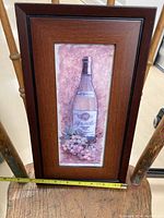 Photo of framed kitchen art print showing a wine bottle with grapes against a pinkish background. Close-up with tape measure for width (approximately 10.25 inches).