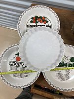 Four pie plates showing three with printed pie labels and decorative rim and one plain ruffled edge plate.