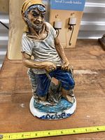 Front view of the Capidimonte crown porcelain figurine of a boy with yellow head covering, light colored shirt, blue pants on water base.