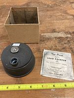 Vintage Tru-Point Model D lead pointer with original cardboard box and abrasive sheet on wood surface with measuring tape.