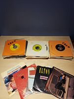 Multiple stacks of 45 RPM vinyl records mostly in orange CBS sleeves, with some visible artist labels and covers like Gloria Estefan and Elton John.