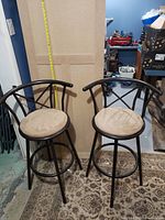 Two black metal bar stools with beige cushioned round seats, X-backrest design, and circular footrest ring, positioned on a patterned rug with measuring tape behind.