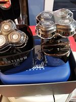 Electric shaver with three rotary heads inside a black rinse stand marked 'Platinum'. The shaver has a shiny metallic finish and a transparent protective cover over the rotary heads, displayed next to its rinse stand.