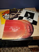 Four comedy vinyl records displayed on a wooden surface: Cheech & Chong "Staging Beauty" with a lip image on the cover, Steve Martin with trumpet face, Richard Pryor "Here and Now" in black and white, and Just for Laughs colorful album cover.