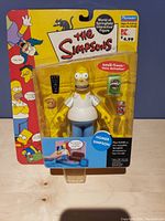 Front view of the Homer Simpson figure in original retail packaging with accessories including a remote control and Duff beer can.
