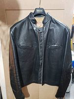 Front view of men's black leather jacket with zipper and snap button closure, two chest pockets, and stand collar.