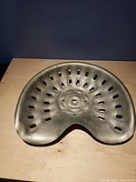Cast metal tractor seat photographed on a wooden surface against dark background showing hole pattern and mounting square hole