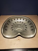 Photo showing the metal tractor seat placed on a wooden surface, highlighting the wide and contoured design with ventilation slots and mounting holes at the center.