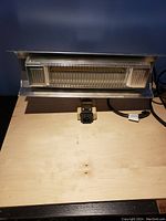 Photo of a rectangular silver metal outdoor electric heater showing the heating element behind a protective grill, power cord and plug connector visible.