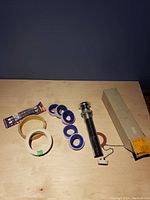 Photo showing all plumbing items on wooden surface: faucet pop-up rod with chain, multiple rolls of blue and white Teflon tape, small packaged item, and boxed pipe fitting.