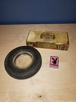 Photo showing a tire-shaped ashtray, a vintage cigar box, and a pink Playboy lighter on a wooden surface in front of a blue background.