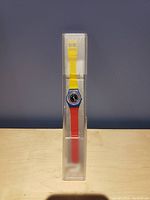 Photo showing new Swatch watch with a yellow and red strap inside clear plastic packaging against plain background.