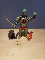 Photo of Mumra action figure holding sword, with Spike figure and red accessory on wooden surface.