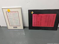 Two framed prints side by side on a floor against a wall. Left frame white with music singles chart and dedication. Right frame black with red cast of characters print.