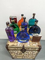 Nine bar decor items consisting of colorful decorative bottles filled with colored water and two metal flasks displayed on a decorative patterned storage box.