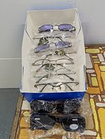 Box with 9 pairs of sunglasses, 6 pairs have clear or purple lenses with mostly metal frames, 3 pairs with dark lenses and plastic frames in bags.