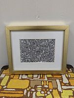 Framed Keith Haring style black and white reproduction print in gold frame sitting against wall on patterned surface.