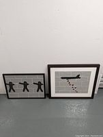 Two framed street art paintings leaning against a white wall, showing full view of both paintings.