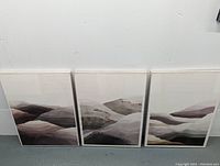 Three framed lithographs leaning against a wall showing abstract layered landscape designs in muted colors.