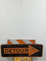 Photo of a rectangular vintage metal road sign with 'DETOUR' in orange letters on a black background, an orange right-pointing arrow, and a top panel with diagonal orange and white stripes, showing signs of wear and rust.