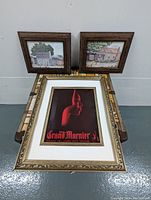 Three framed prints arranged on a patterned surface: two smaller folk art prints and one larger Grand Marnier promo print with decorative gold frame and white matting.