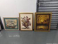 Lot of three framed vintage artworks displayed leaning against a white wall and grey floor.