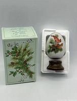 Porcelain egg with bird design displayed in its original foam packaging alongside the original box showing product details and winter themed foliage illustration.