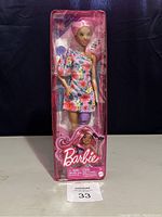 Barbie Fashionistas Doll #189 in box, showing pink hair, floral one-shoulder dress, and purple prosthetic leg.