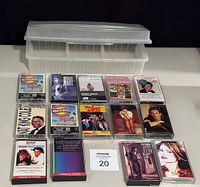 Lot of 14 assorted music cassette tapes arranged in rows in front of a clear plastic storage box with lid.