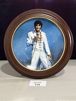 Frontal image of the collectors plate showing Elvis Presley in a white jumpsuit holding a microphone, framed in a wooden circular frame with a gold trimmed edge.
