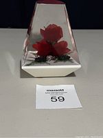 Triangular clear resin paperweight containing red roses and small foliage with a white base on a gray surface
