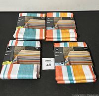 Photo showing all 4 folded tablecloths with visible packaging labeled Mainstays, price tags, and striped pattern with blue, orange, white, and yellow colors.
