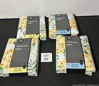 Four unopened tablecloths displayed with floral yellow flower and green leaf pattern and visible packaging size labels.