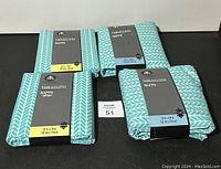 Four packaged tablecloths with teal and white herringbone patterns laying flat on a black surface.